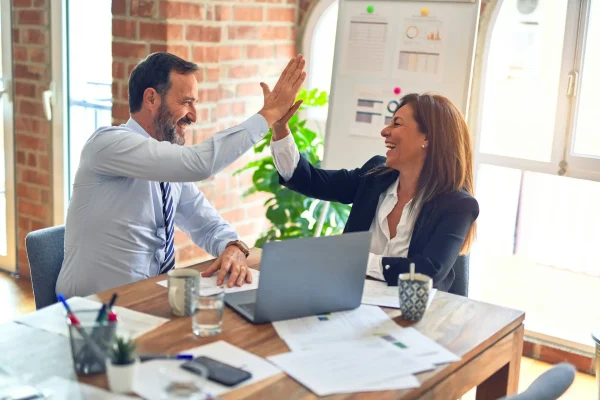Business professionals smiling and high-fiving at a desk with laptops, papers, and coffee, representing teamwork and success.
