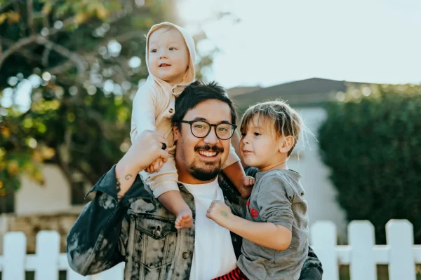 A smiling father with glasses holding his two young children outdoors, standing in front of a white picket fence with soft sunlight in the background.